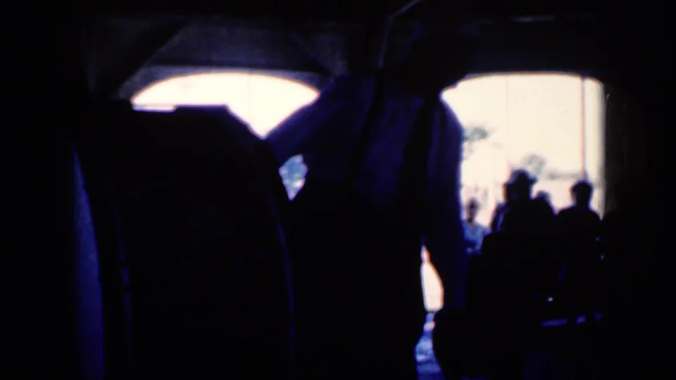 1959 Iowa 8mm frame showing a dim hall-like interior, audience silhouettes, and a white-shirted performer near bright arched window openings.