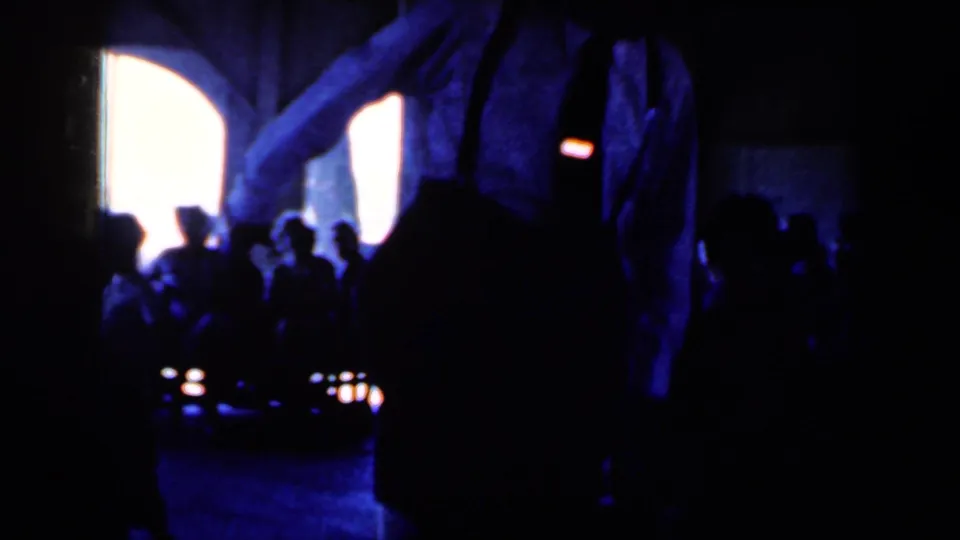 Dim 1959 Iowa home-movie frame with clustered audience silhouettes, arched bright windows, and the performance area largely in shadow.
