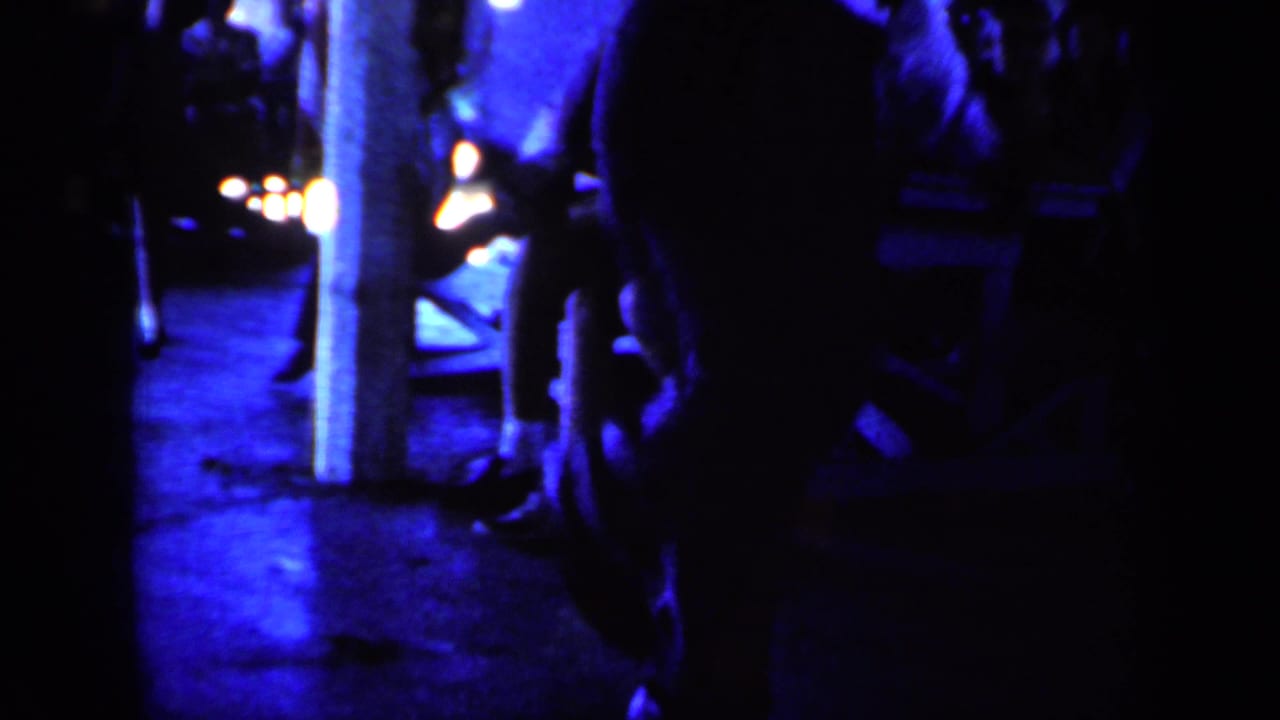 Dim blue-lit room with a man in a white shirt partly visible near posts and background figures.