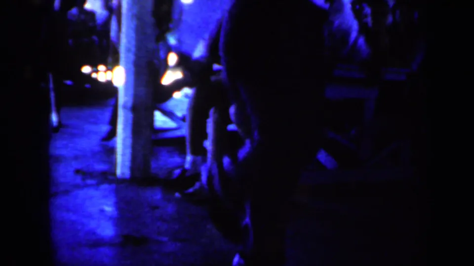 1959 Iowa 8mm frame showing a dim blue-lit interior with a man in a white shirt moving near posts and indistinct background figures.