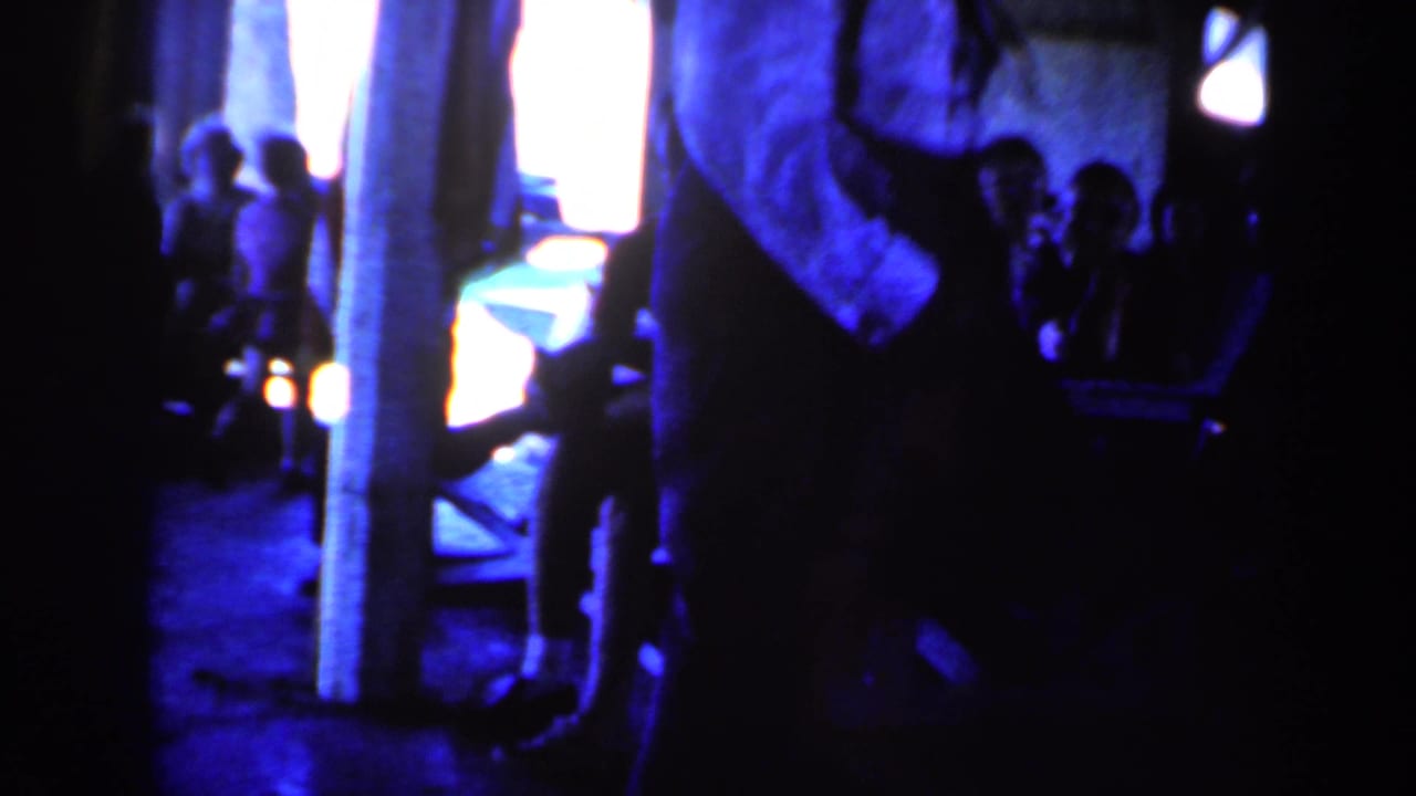 1959 Iowa home-movie frame with saturated blue light, vertical posts, indistinct people, and the dancer partly obscured at right.