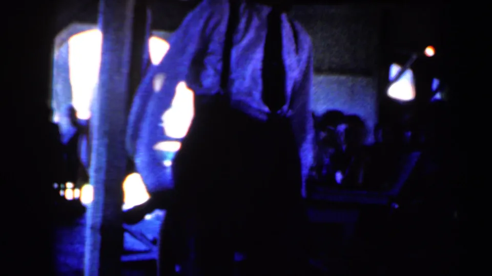 1959 Iowa frame showing a man in a white shirt and dark trousers dancing energetically in a dim, blue-lit indoor space.