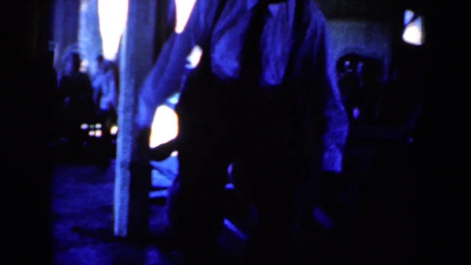1959 Iowa 8mm frame with the dancer near a vertical post, surrounded by deep shadows, blue light, and indistinct bystanders.