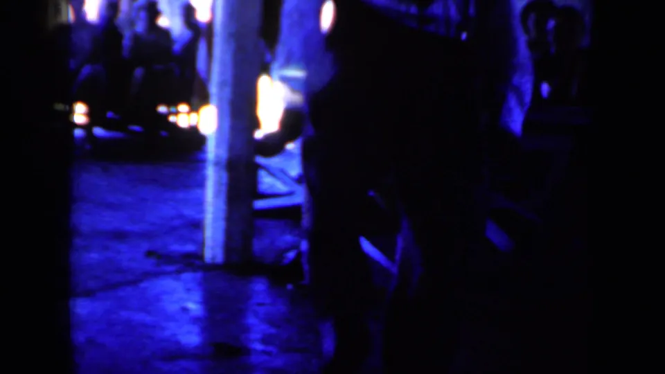 1959 Iowa home-movie frame showing silhouetted people on a dim blue train platform with bright station lights behind them.