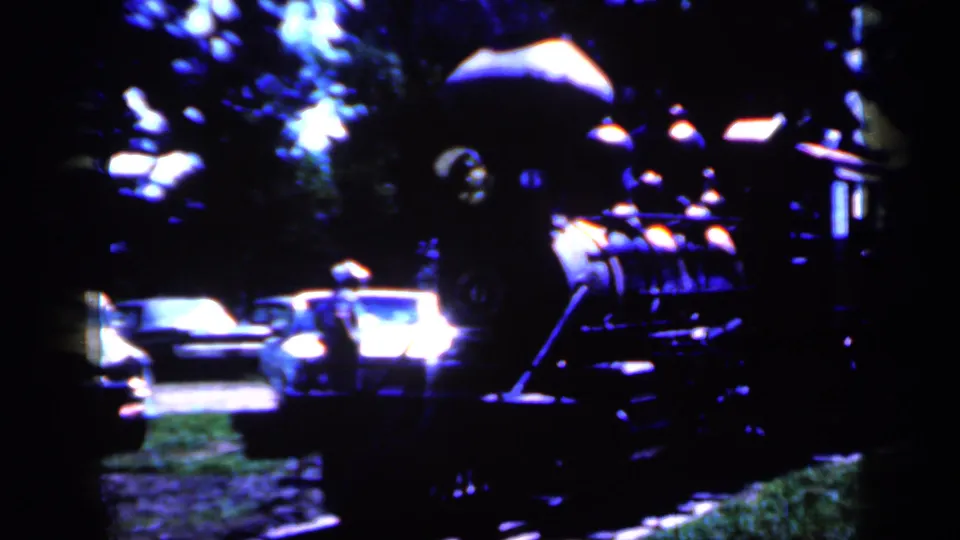 1959 Iowa home-movie frame with a steam-style train form or model train beside parked cars, trees, and deep blue shadows.