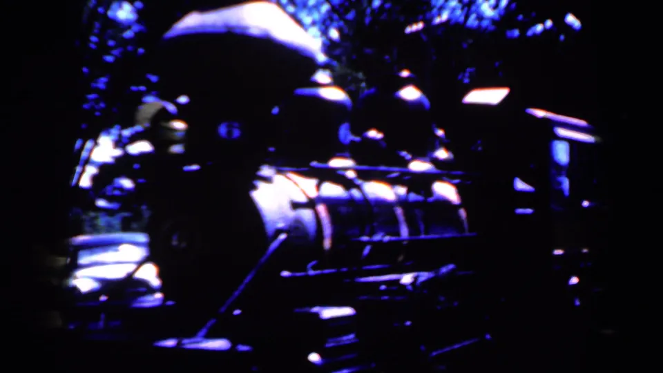 Blue-shifted 1959 Iowa frame showing a train-like engine form in shadow, with nearby cars and trees partly visible.