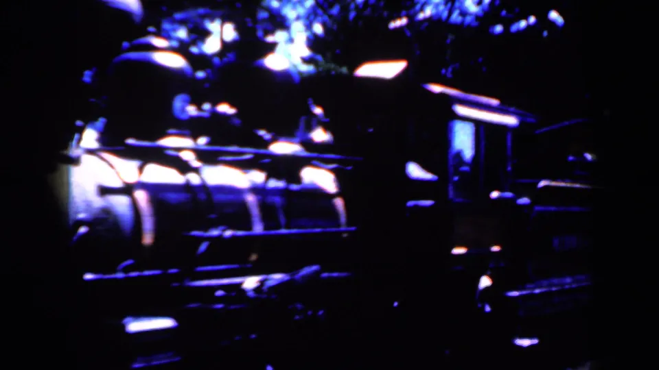 1959 Iowa home-movie frame showing a dark locomotive at night with bright lamp reflections and wooded background.