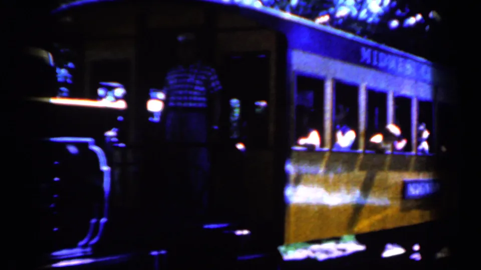 A 1959 Iowa passenger car carries visible riders while a man in a plaid shirt stands near the train at night.