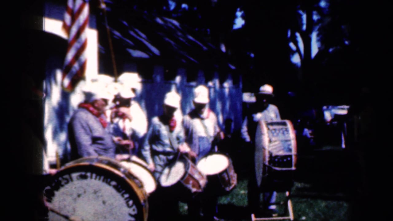 Band members in white hats and blue uniforms gather with drums under trees near an American flag in 1959 Iowa fair footage.