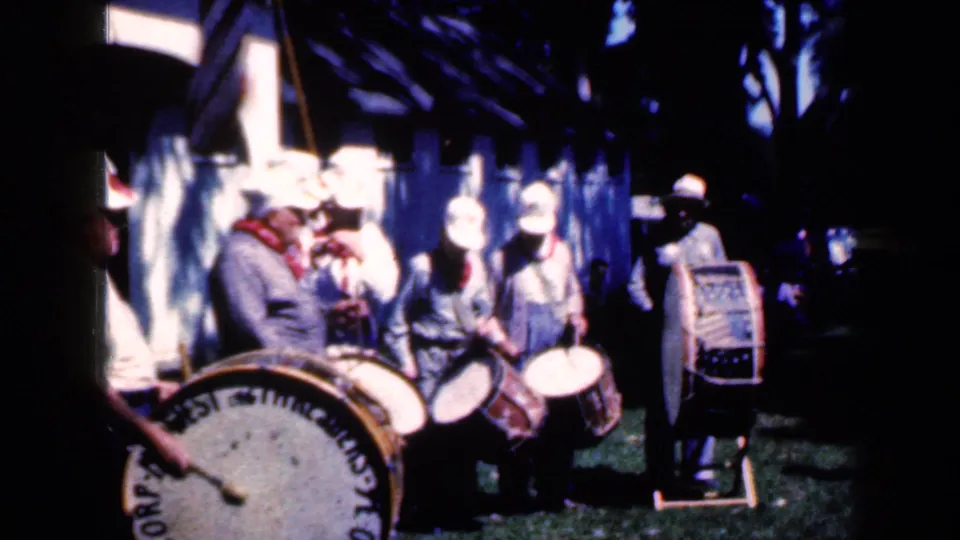 At the Iowa fair, uniformed drummers with white hats stand in shade with a large bass drum partly filling the foreground.