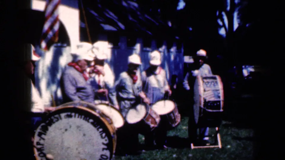 Uniformed band members with drums stand near an American flag and shaded trees during the 1959 Iowa fair scene.