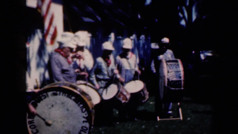 1959 Iowa home-movie frame showing uniformed drummers with a large “CORPS OF DRUMS” bass drum and U.S. flag behind them.