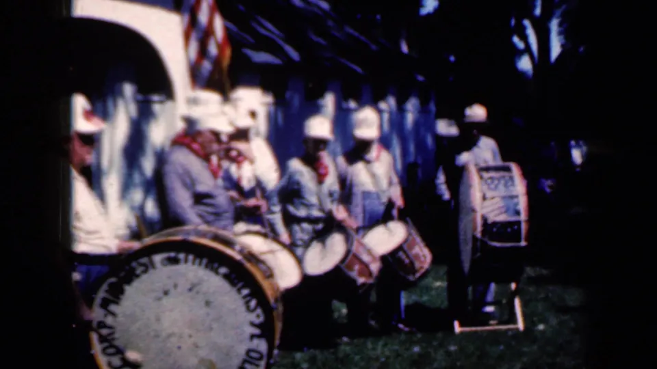 1959 Iowa home-movie frame showing uniformed drummers, a foreground bass drum, and a spectator or participant entering at the left edge.