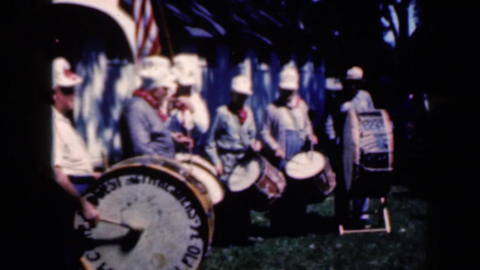 1959 Iowa frame showing a compact drum corps group performing outdoors, with the bass drum lettering and U.S. flag still partly visible.