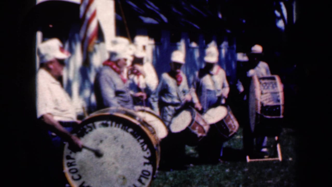 Uniformed parade musicians with drums stand near arches and an American flag.