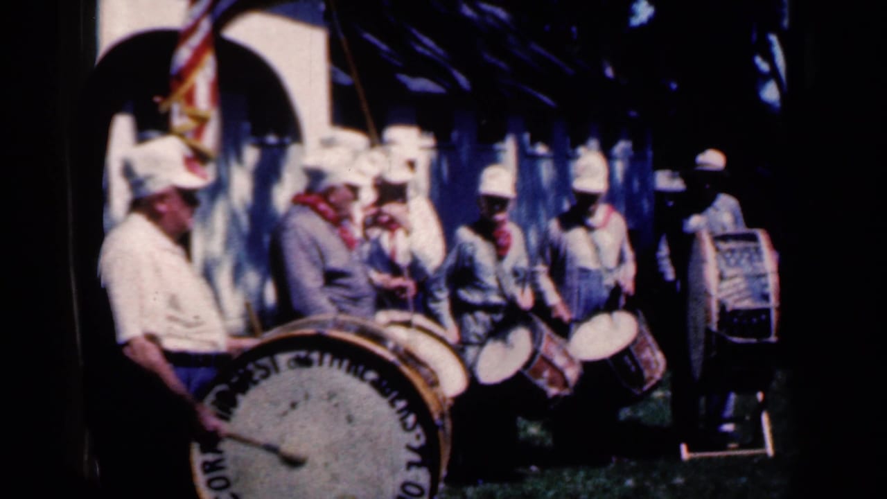 Marching band members play drums with a flag and tuba visible behind them.