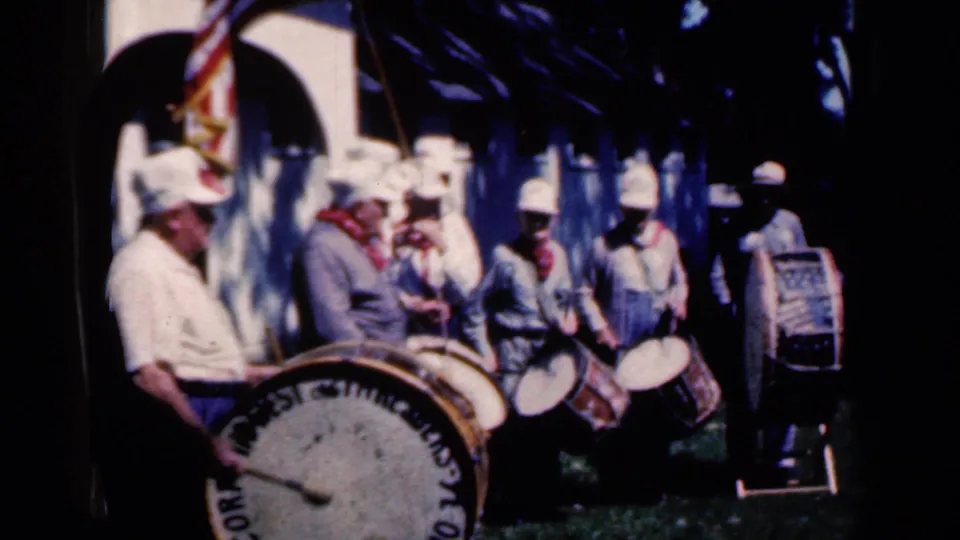 1959 Iowa parade footage frame with drummers in light uniforms, a tuba player, and an American flag in a public outdoor setting.