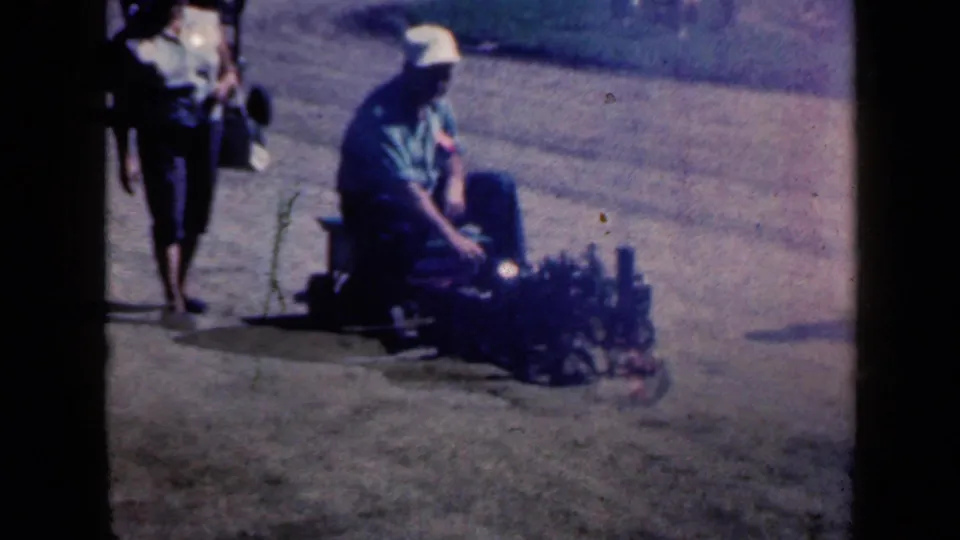 Home-movie frame from the 1959 Iowa clip showing a person seated on a small parade vehicle or float, with another pedestrian nearby.