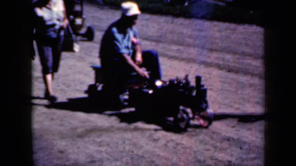 1959 Iowa home-movie frame showing a seated participant on a small parade vehicle or float, filmed from the side on a paved street.