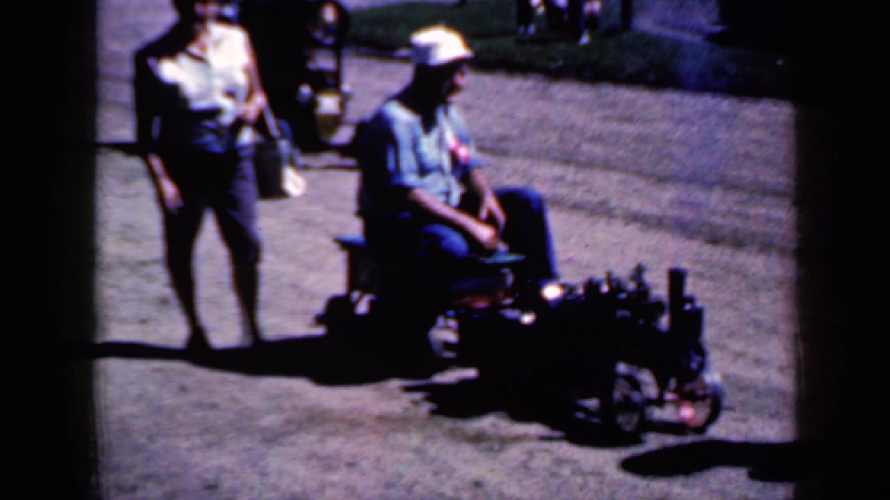 Woman walks beside a seated man riding a small motorized vehicle on grass near a paved path.