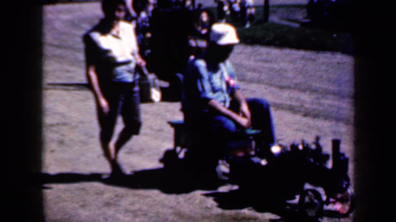 The man continues riding the small motorized toy vehicle as the woman keeps pace in the 1959 Iowa outdoor scene.