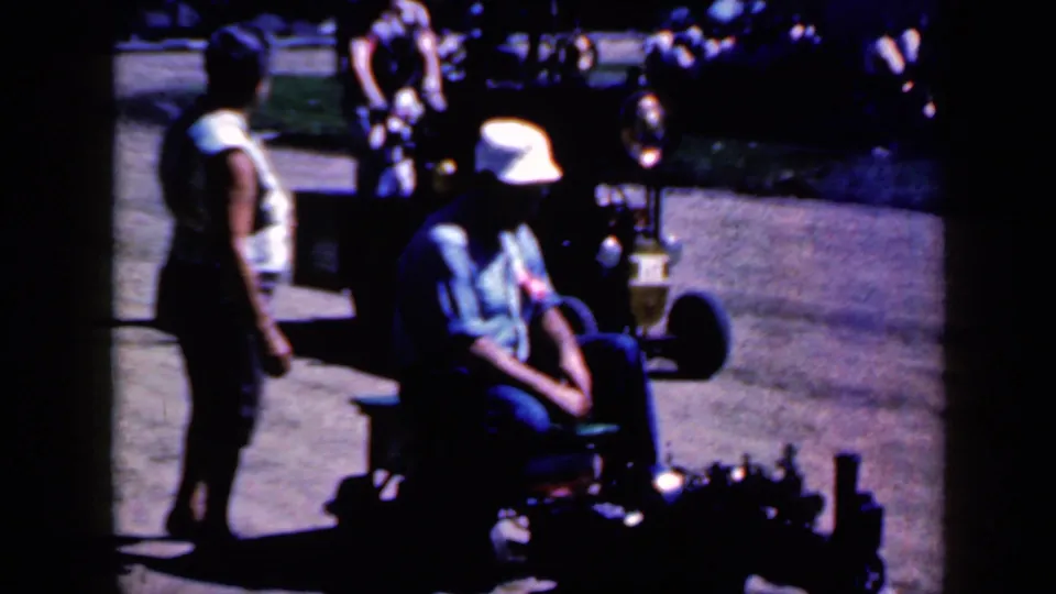 A woman stands close to the seated rider on the small motorized vehicle, with grass and walkway visible in the Iowa home movie frame.
