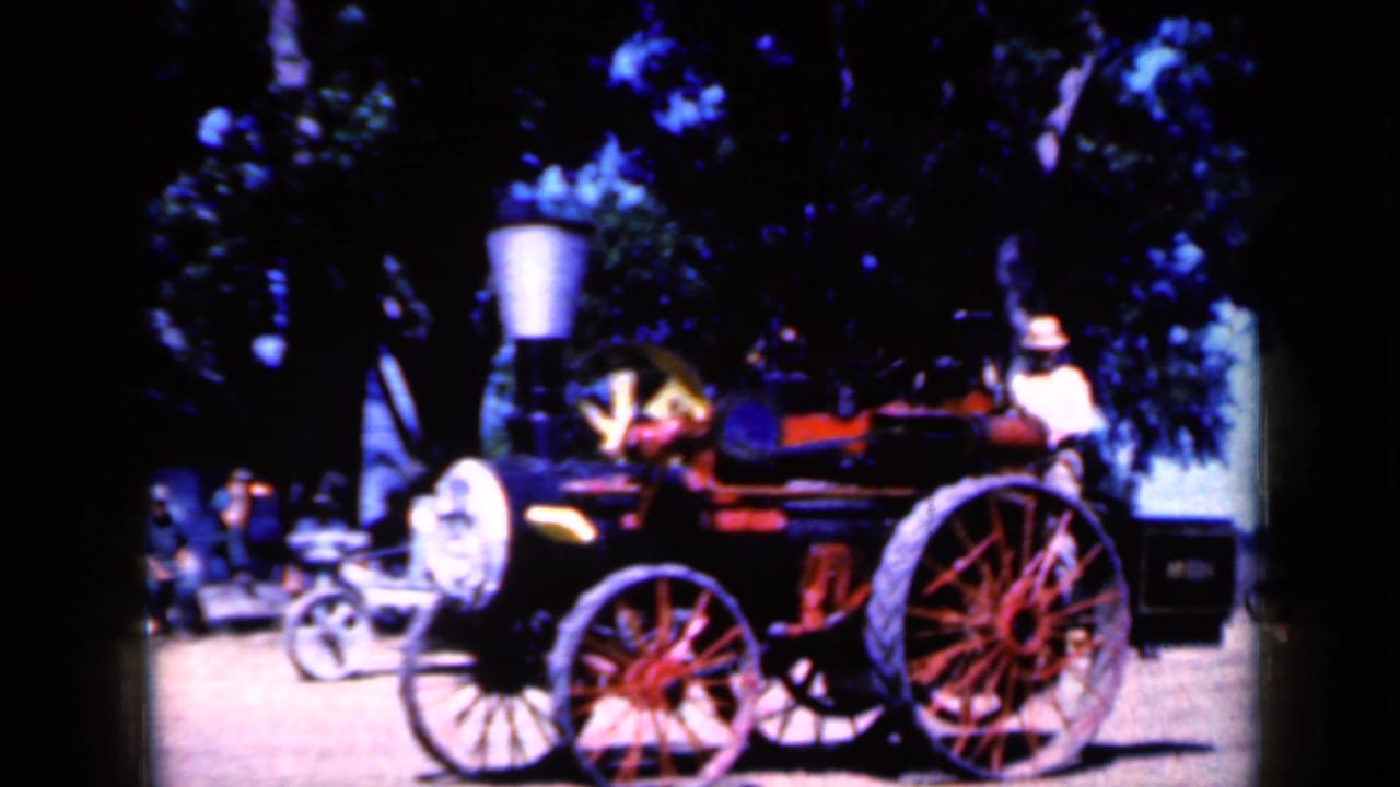In Iowa 1959 footage, an adult man operates a small steam-powered vehicle with large spoked wheels among trees.