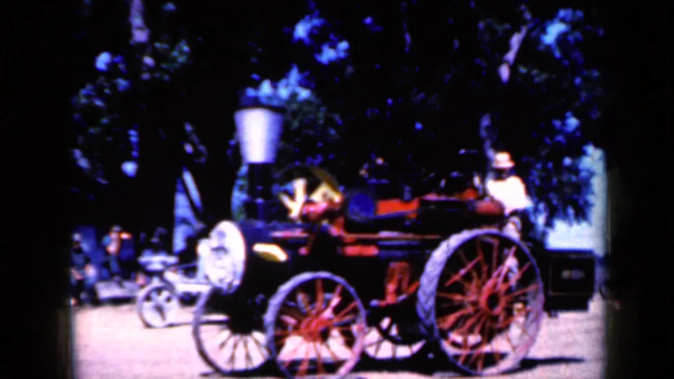 In Iowa 1959 footage, an adult man operates a small steam-powered vehicle with large spoked wheels among trees.
