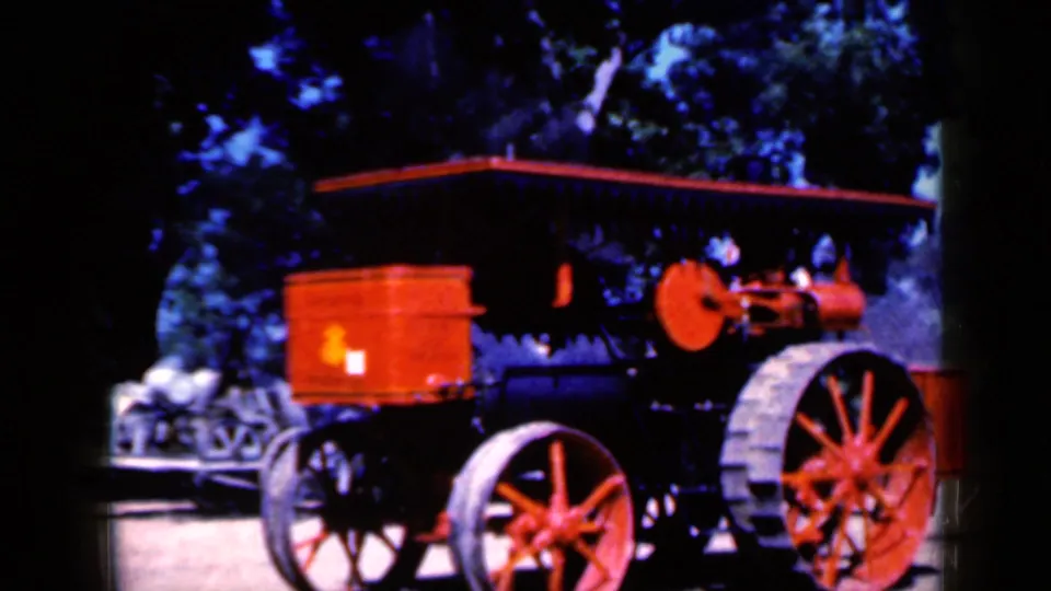 1959 Iowa home-movie frame with the red and black steam tractor filling the view, framed by trees and bright daylight.