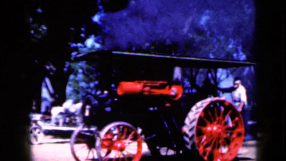 A side view shows the red and black steam vehicle, large red rear wheel, smoke or steam, trees, and nearby display equipment.