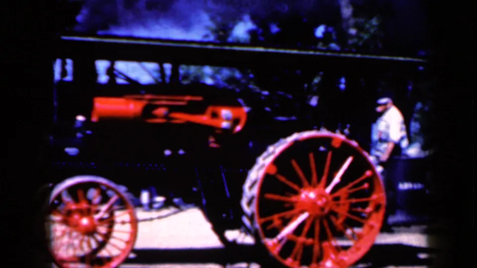 1959 Iowa home-movie frame with a closer side view of the steam tractor, red wheels, black body, and a man beside it.