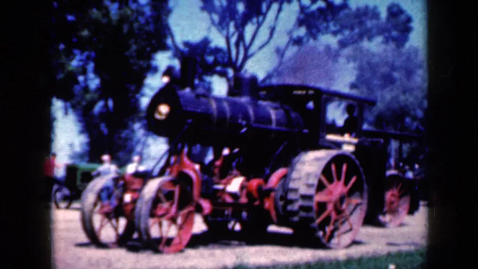 The steam tractor is seen from the front side in the 1959 Iowa footage, with a dark boiler, red wheels, headlamp, and trees behind it.