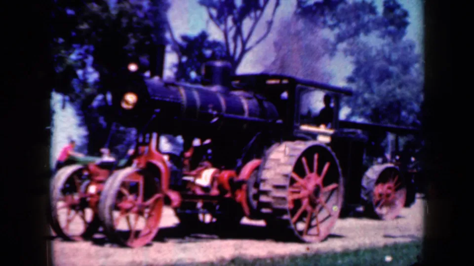 1959 Iowa home-movie frame of a black steam tractor with red spoked wheels, headlamp and cab on a tree-lined dirt path.