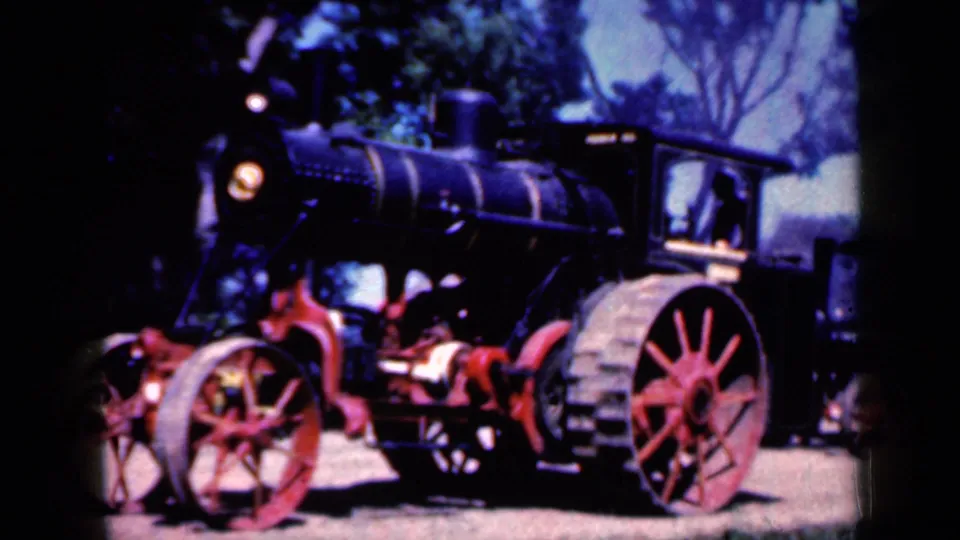 Close 1959 Iowa view of a steam tractor showing the black boiler, red spoked wheels and mechanical parts under trees.