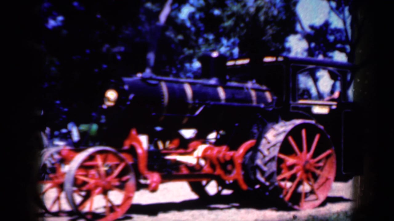 Side view of the steam-powered tractor in Iowa, with a dark boiler, red running gear, and trees behind it.
