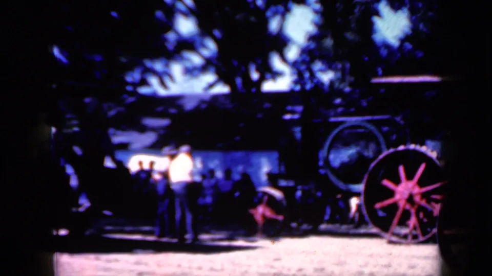 Wider 1959 Iowa home-movie view with onlookers standing near the steam tractor in a shaded outdoor display area.