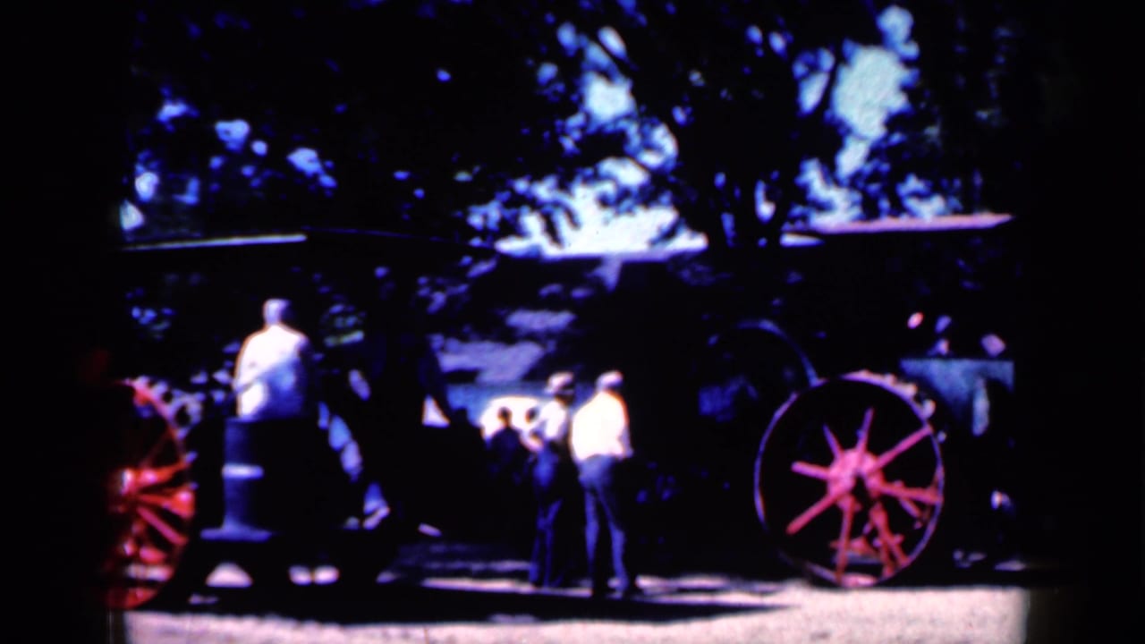 Men and other spectators in Iowa appear to watch the old steam tractor as it moves through a shaded outdoor area.