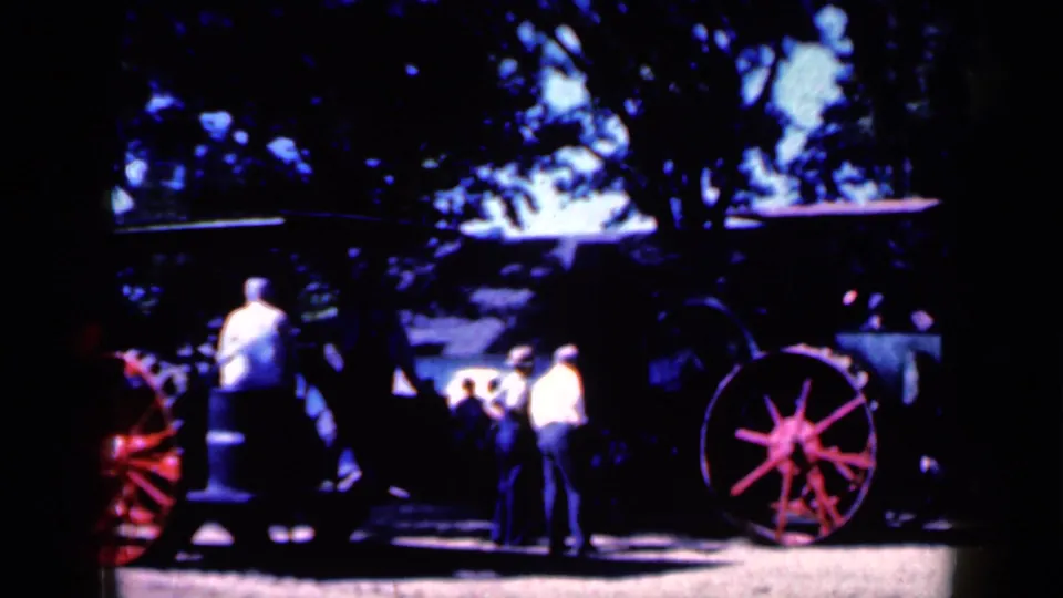 Men and other spectators in Iowa appear to watch the old steam tractor as it moves through a shaded outdoor area.