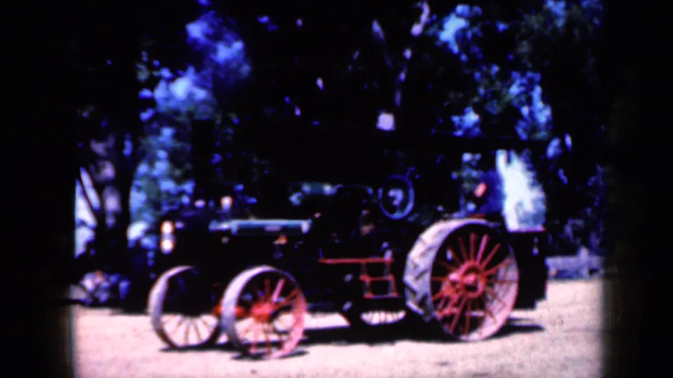 The old steam tractor continues past on a dirt path, framed by dark tree shade and bright home-movie color shifts.