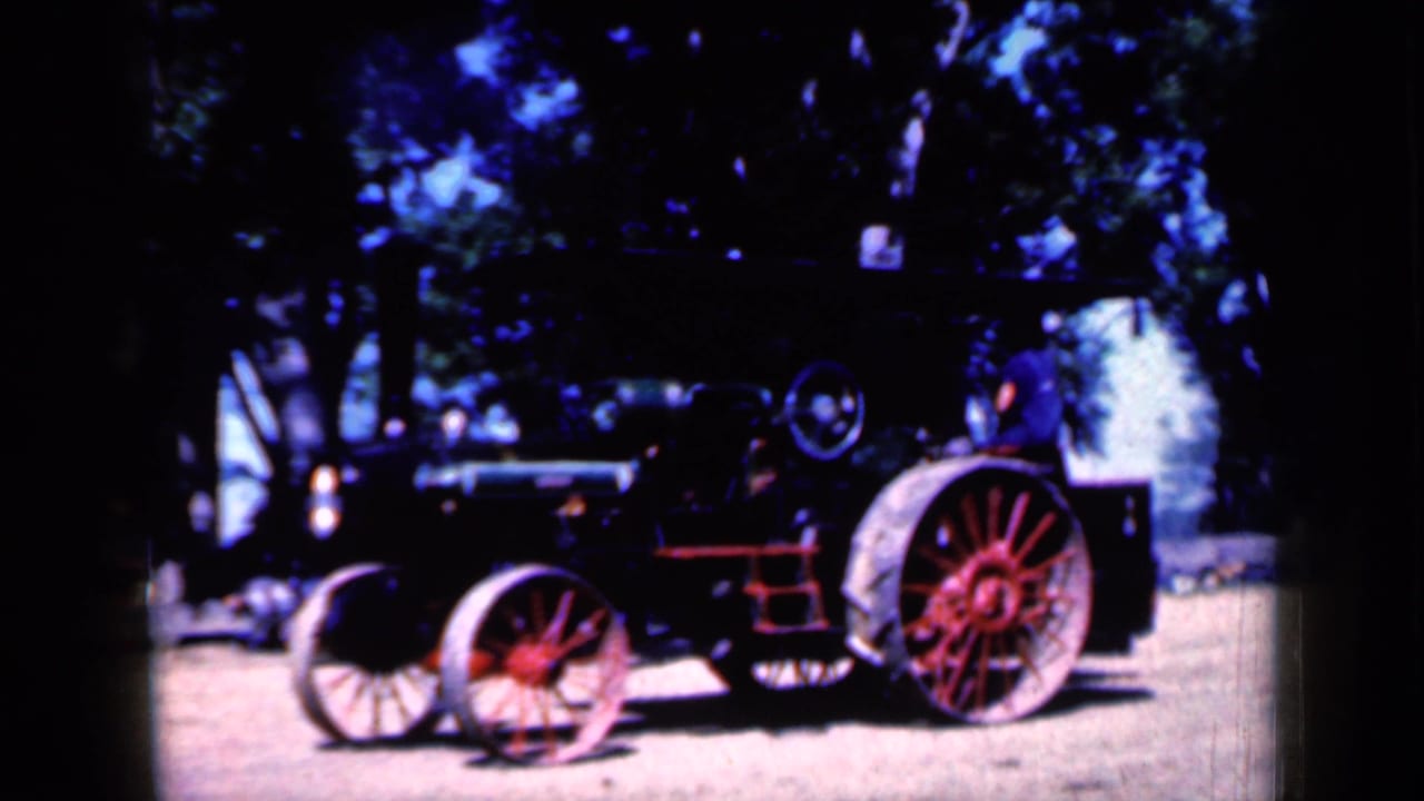 Red and black steam tractor with large spoked wheels parked on dirt under trees.