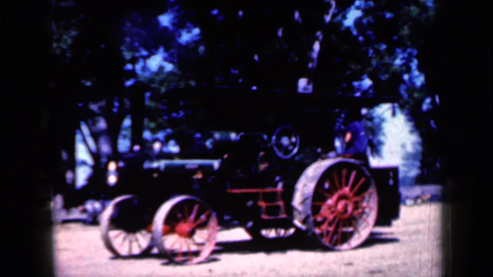 1959 Iowa home-movie frame showing a red and black steam-powered tractor with spoked wheels and a tall stack under leafy trees.