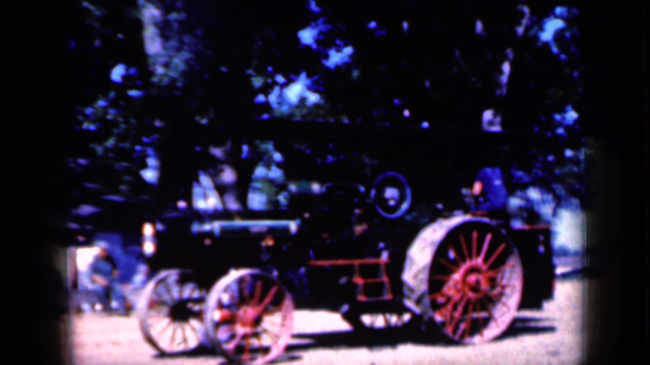 Steam tractor with red wheels and a person nearby in a shaded outdoor display area.