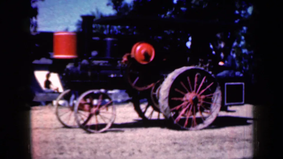 Closer 1959 Iowa frame of the steam tractor, emphasizing the boiler, red smokestack cap, front wheel assembly, and large spoked rear wheel.
