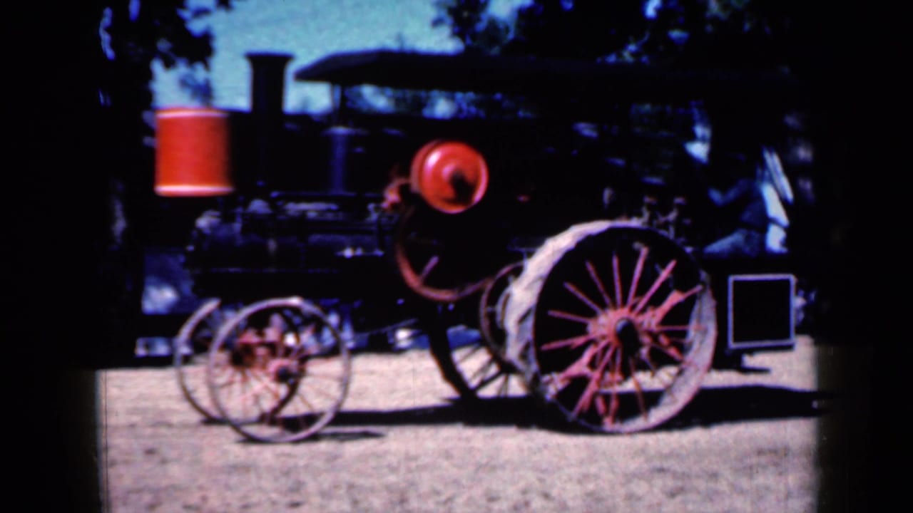 Antique steam tractor with red spoked wheels standing on dirt beneath trees.