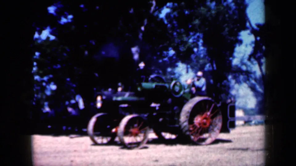 1959 Iowa frame showing the antique steam tractor from a wider angle, with trees, dirt ground, and a person briefly visible nearby.