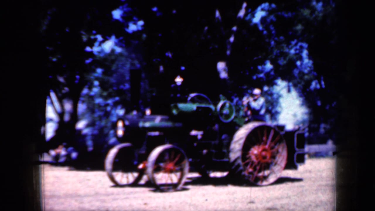 1959 Iowa home-movie view of a steam tractor with red wheels, smoke, and a seated operator in a light hat.
