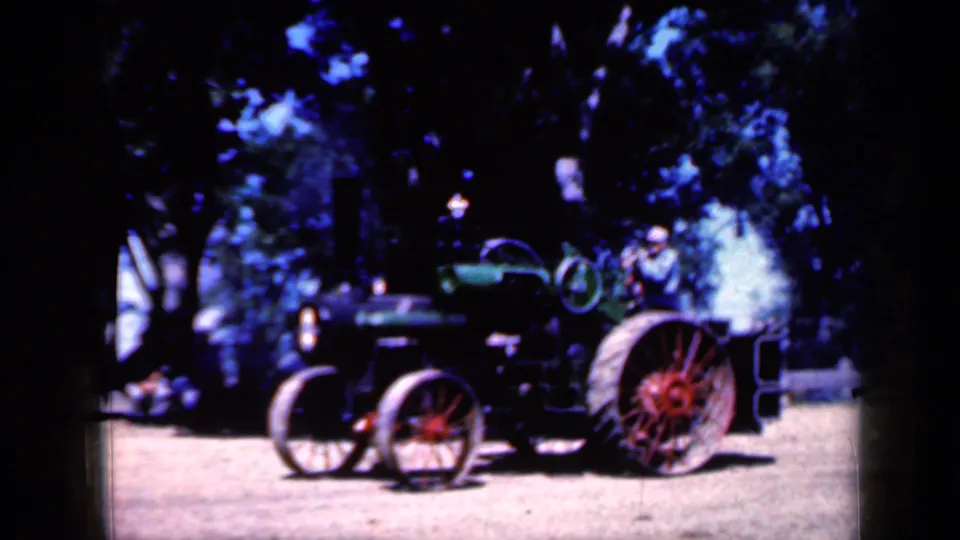 1959 Iowa home-movie view of a steam tractor with red wheels, smoke, and a seated operator in a light hat.