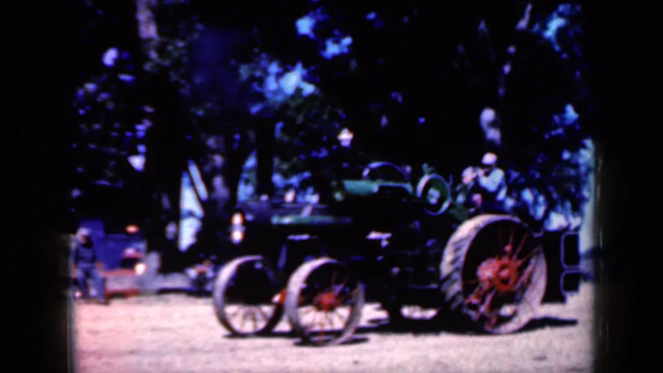 1959 Iowa frame showing the steam tractor under trees, with red wheels and small background figures near the left edge.