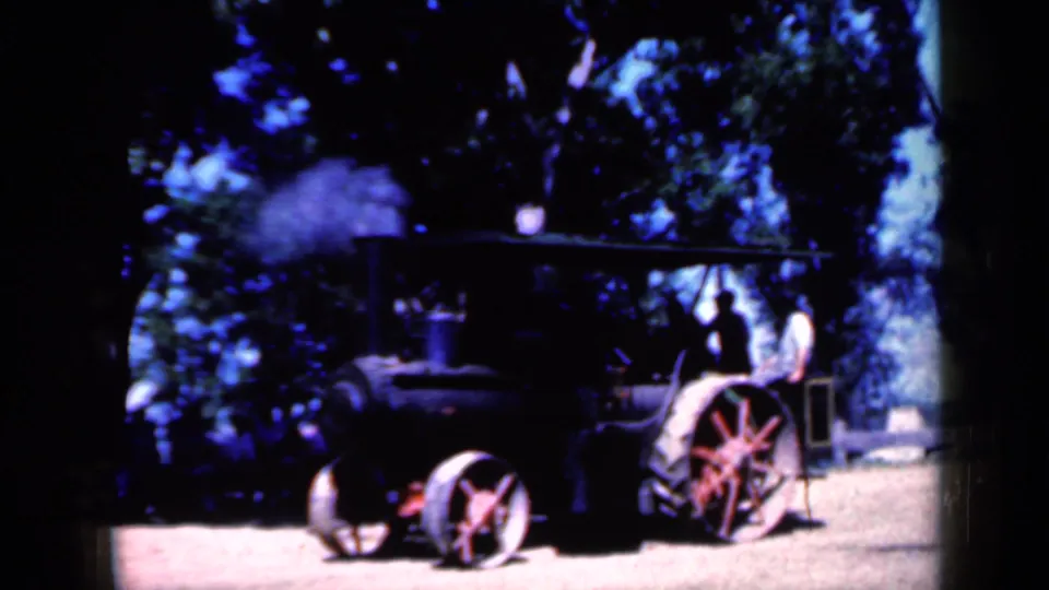 1959 Iowa home-movie frame of a steam tractor in side view, with smoke rising and a few people near the rear.