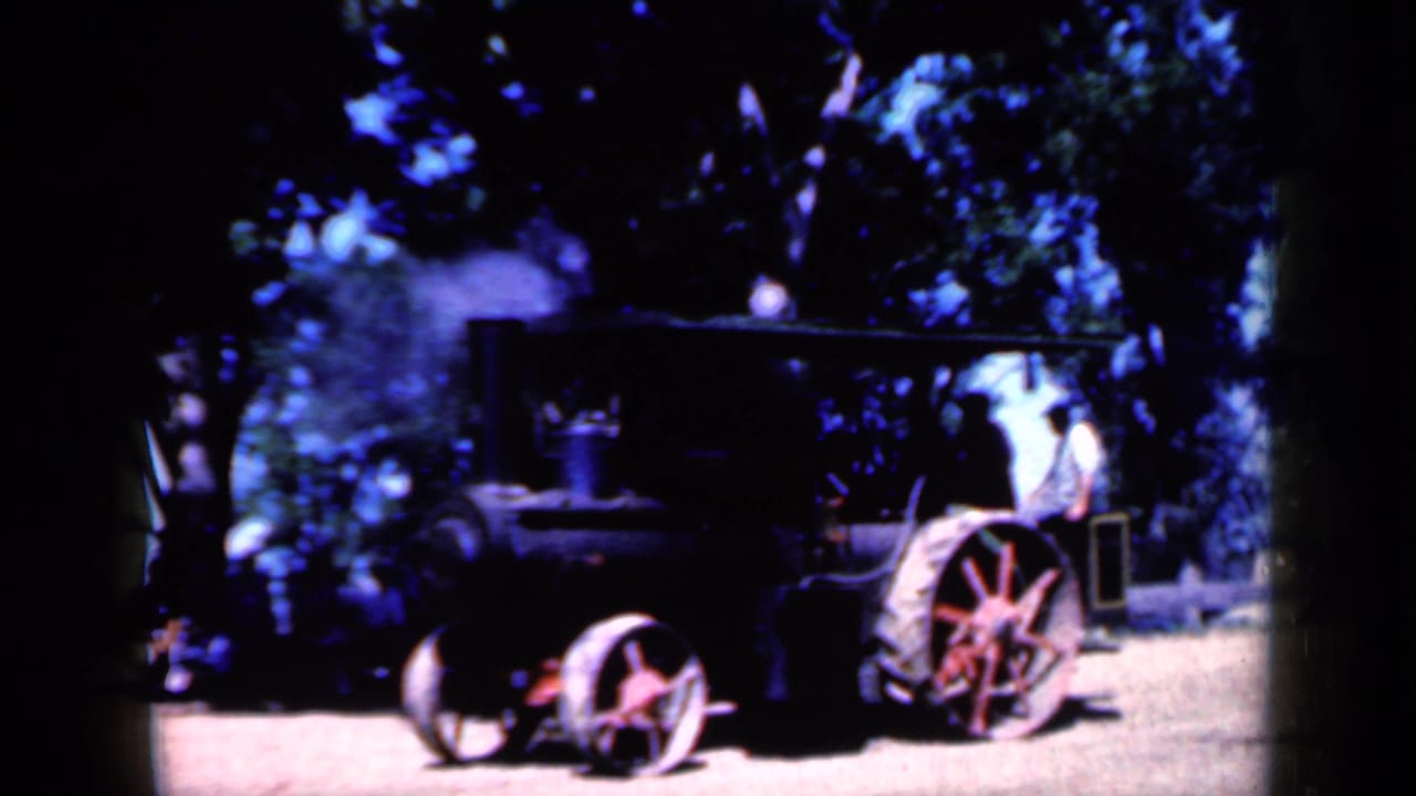 Black steam tractor with red wheels on dirt under trees, with steam rising nearby.